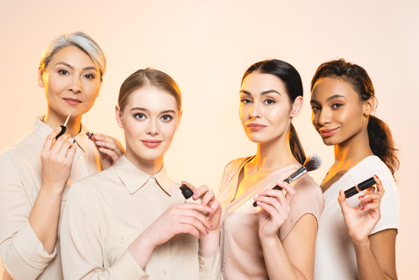 happy multicultural women holding decorative cosmetics isolated on beige  - Photo, Image