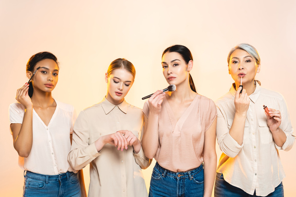 atractivas mujeres multiculturales que sostienen cosméticos decorativos y cepillos cosméticos aislados en beige
  - Foto, imagen