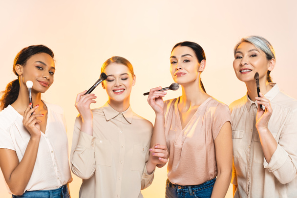 happy multicultural women holding cosmetic brushes isolated on beige  - Foto, imagen