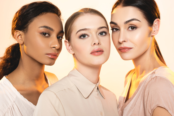 three beautiful multicultural women with makeup looking at camera isolated on beige  - Photo, Image