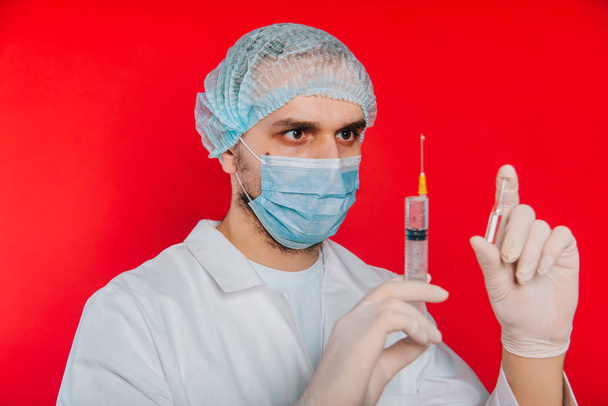 Lékař drží injekční stříkačku a ampulku s vakcínou. Mladý muž v bílém plášti, v lékařské masce a rukavicích na červeném pozadí. - Fotografie, Obrázek