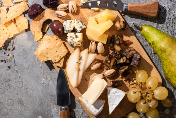 Vue de dessus de différents types de fromage aux olives séchées, pistaches, raisins, poires et craquelins sur planche à découper
  - Photo, image
