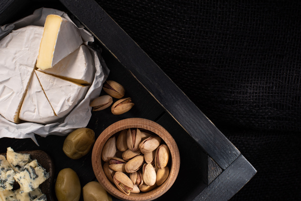 Top view of tray with pieces of camembert and dorblu, dried olives and pistachios isolated on black  - Fotografie, Obrázek