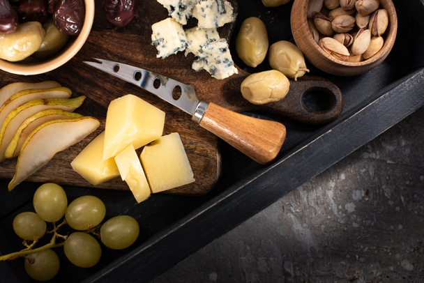 Top view of tray with pieces of cheese, dried olives, pistachios, slices of pear, grapes and knife on grey background - Foto, Bild