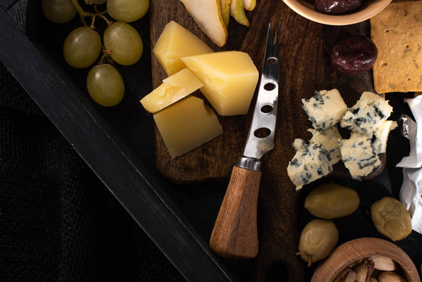 Top view of tray with knife next to pieces of cheese, slices of pear, olives, pistachios and grapes isolated on black  - Foto, imagen