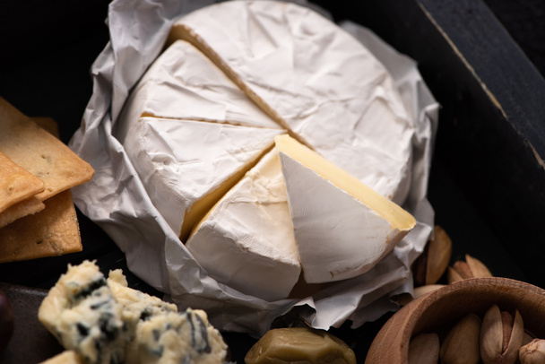 Selective focus of pieces of camembert with crackers, bowl of pistachios, dorblu and olive on tray - Photo, Image