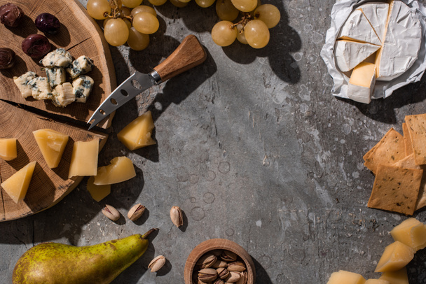 Top view of cheese, fruits, nuts, crackers and olives with knife and cutting board on grey background - Photo, Image