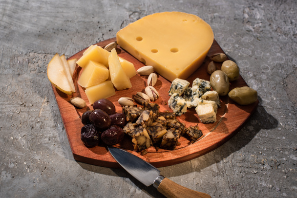 Nahrungsmittelzusammensetzung aus Käse mit getrockneten Oliven, Birnenstücken, Pistazien und Messer auf Schneidebrett auf grauem Hintergrund - Foto, Bild