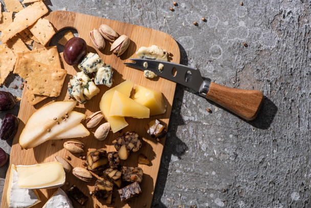 Top view of pieces of cheese, olives, pistachios, slices of pear, crackers and knife on cutting board on grey background - Photo, Image