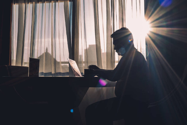 La silhouette d'un homme au travail à l'aide d'un ordinateur portable dans une pièce moderne avec un fond de fenêtre éclairé par des reflets naturels du soleil légèrement éblouissants
. - Photo, image