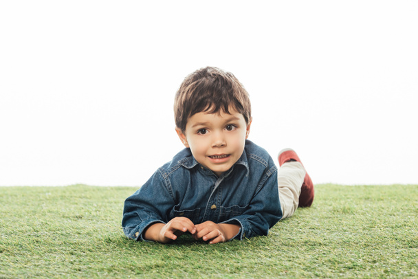 bonito e sorridente menino olhando para longe e deitado na grama isolado no branco
  - Foto, Imagem
