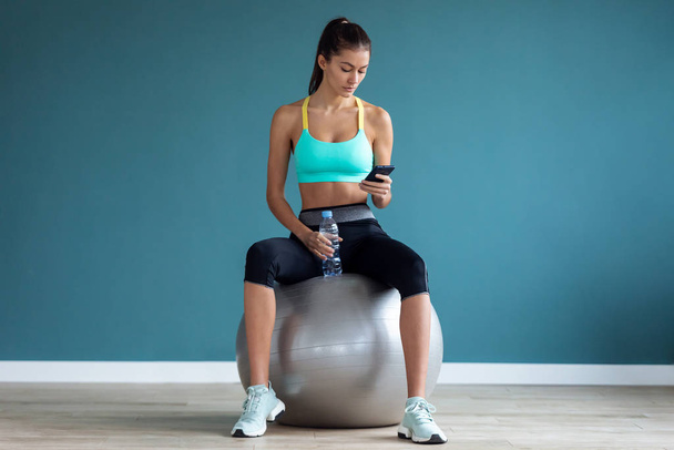 Sporty young woman using her mobile phone while holding water bottle after a pilates class on the gym. - Foto, Imagem