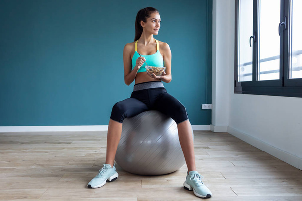 Sporty young woman eating a bowl of muesli while sitting on fitness ball at home. - Foto, Bild