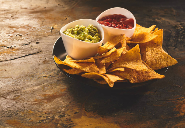 Single serving of delicious crunchy appetizer of triangular yellow corn tortilla chips beside little bowls of guacamole and salsa dip - Photo, image
