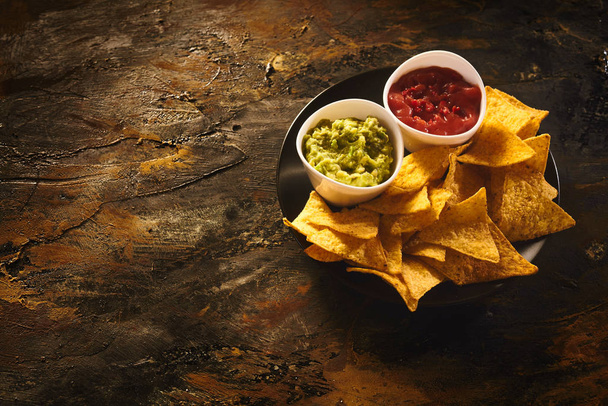 Top down view on delicious crunchy appetizer of triangular yellow corn tortilla chips beside little bowls of guacamole and salsa dip over old dark wooden table with copy space - Photo, Image