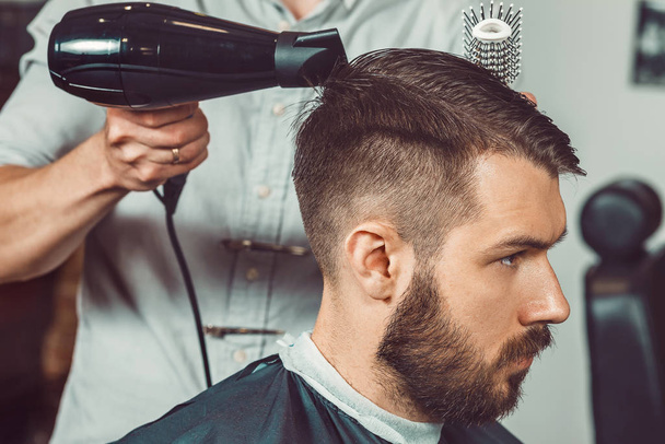 The hands of young barber making haircut of attractive bearded man in barbershop - Foto, Imagen