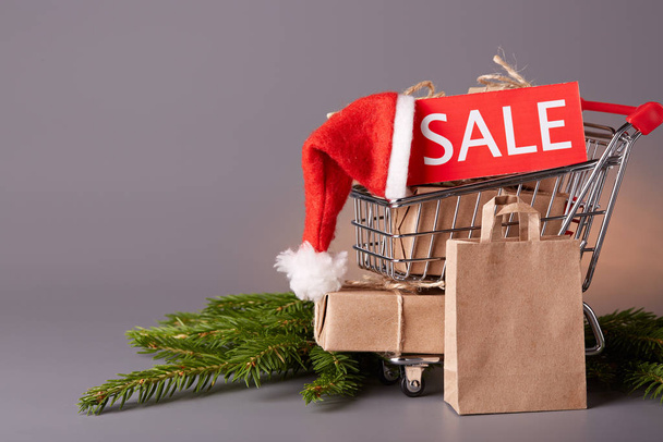 Shopping trolley with gift boxes, tablet with an inscription sale and brenches of fir-tree on a grey background. Christmas and New Year sale. - Zdjęcie, obraz