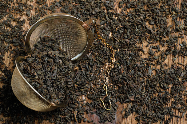 Μαύρο τσάι σε σουρωτήρι με αλυσίδα σε διάσπαρτα τσάι σε ξύλινο φόντο. - Φωτογραφία, εικόνα