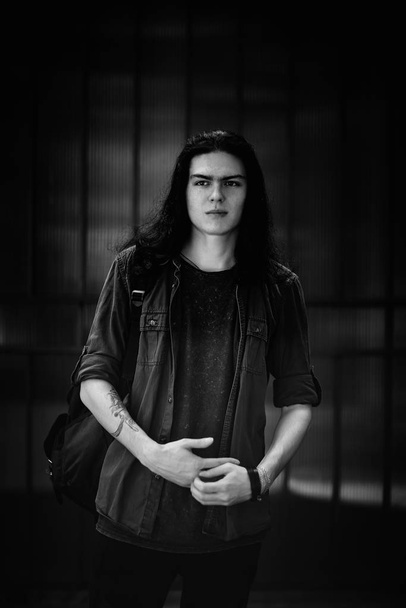 a black-and-white photo of a young man with long, dark hair and brown eyes, a face with freckles, walking down the street in a stylish shirt and jeans,in a Cagle style, in a sad mood - Fotografie, Obrázek