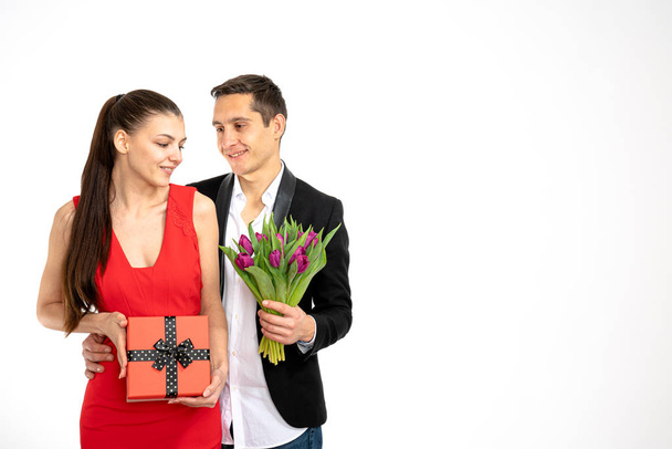 Beautiful romantic couple isolated on white background. Attractive young woman in red dress and handsome man in white shirt are standing with gift box and purple tulips in hands. Happy Saint Valentine - Fotografie, Obrázek