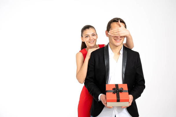 Valentine Gift. Happy young couple in elegant clothes with Valentines Day present isolated on a white background. Happy brunette girl in red dress giving a gift to her boyfriend in black blaser - Foto, immagini