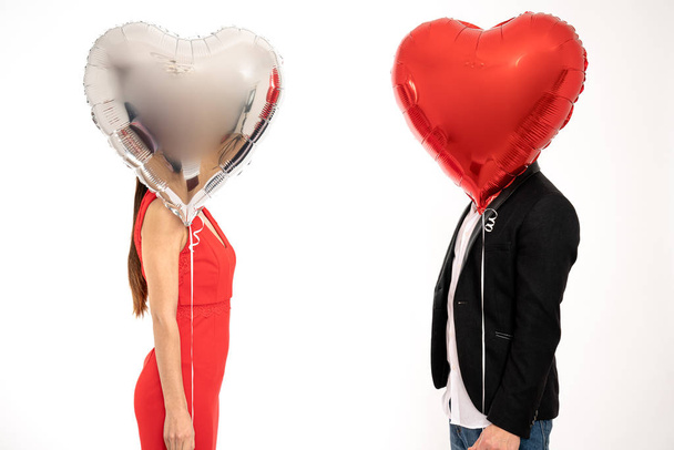 Elegant couple stands against each other, covering their faces with silver and red air balloons, St. Valentines day concept, isolated over white background - Foto, Imagen