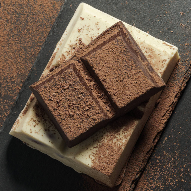 Barres de chocolat blanc et noir empilées sur fond de pierre brute noire recouverte de poudre de cacao
 - Photo, image