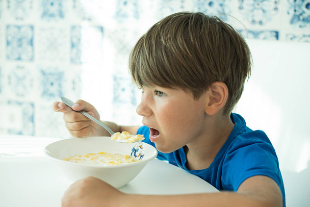 chłopiec w niebieskiej koszulce je śniadanie z owsianką i mlekiem w białym talerzu, miejsce na tekst, izolat - Zdjęcie, obraz