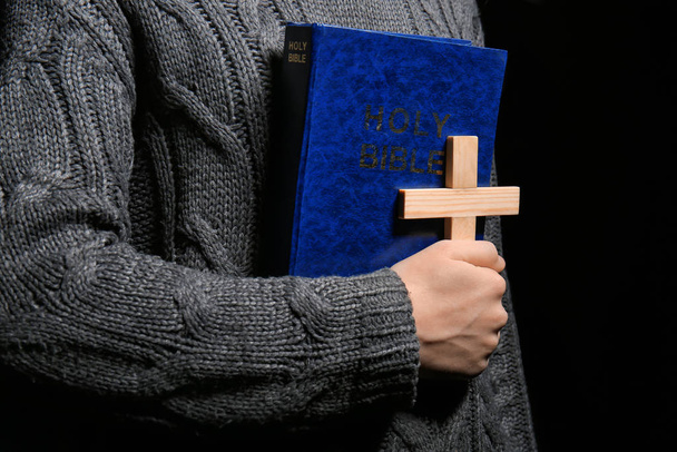 Man with Bible and cross on dark background, closeup - Photo, Image