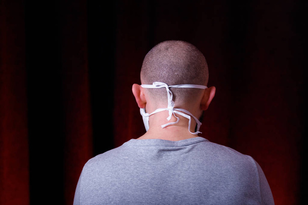 Mies lääketieteellisessä sideharso naamio seisoo takaisin kameran tummanpunainen tausta. Virusten ennaltaehkäisymenetelmät. Ylä- ja alahengityselinten suojaaminen kiinalaiselta koronavirukselta
. - Valokuva, kuva