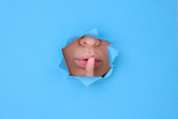 Part female face lips and tongue of attractive woman, girl covers her lips with her finger symbolizes silence behind broken wall or torn paper, blue background. Space for text, feminist banner - 写真・画像