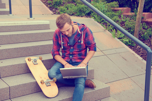 young man in checkered shirt sitting on stairs and using laptop - Valokuva, kuva