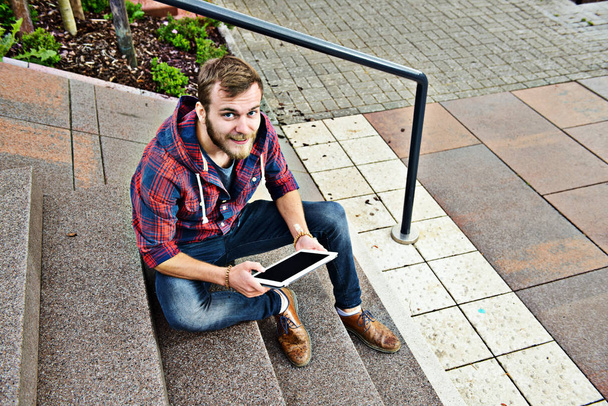 young man in checkered shirt sitting on stairs and using tablet PC - Fotoğraf, Görsel