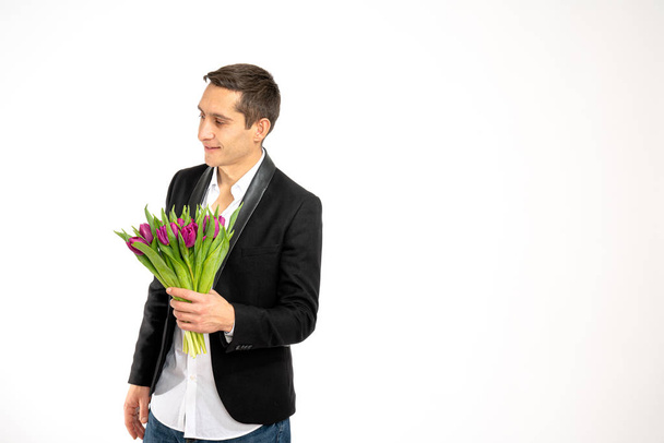 Attractive young man in black blaser holds bouquet of purple tulips in his hand and looking at left side isolated over white background with copyspace - Photo, Image