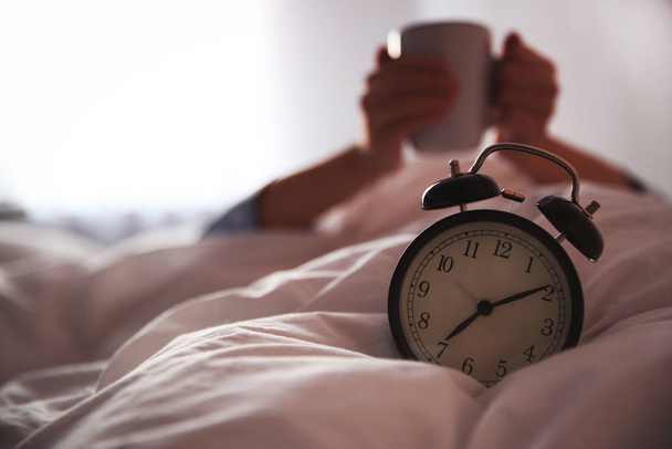 Woman with cup in bed, focus on alarm clock. Morning time - Photo, Image