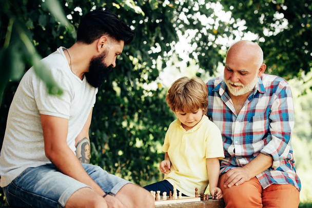 Cute little boy playing chess with Parents. Three different generations ages: grandfather father and son together. - Zdjęcie, obraz