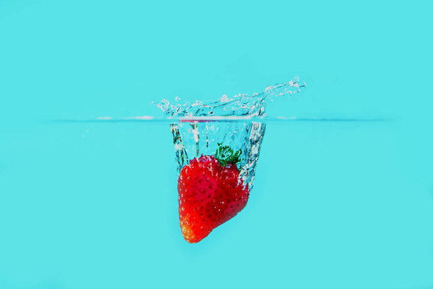 Μια φρέσκια και νόστιμη κόκκινη φράουλα απομονωμένη σε μπλε φόντο. Κόκκινη φράουλα πέφτει στο νερό και δημιουργεί μια βουτιά. Η έννοια της υγιεινής διατροφής, της κατανάλωσης φρούτων. - Φωτογραφία, εικόνα
