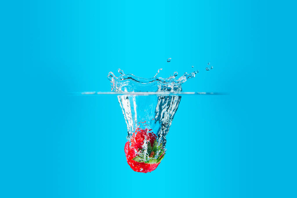 Μια φρέσκια και νόστιμη κόκκινη φράουλα απομονωμένη σε μπλε φόντο. Κόκκινη φράουλα πέφτει στο νερό και δημιουργεί μια βουτιά. Η έννοια της υγιεινής διατροφής, της κατανάλωσης φρούτων. - Φωτογραφία, εικόνα