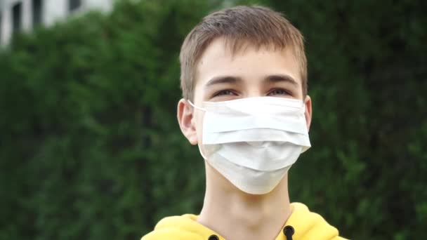 Fiatal férfi tinédzser egy védő orvosi kék maszkban nevet a zöld növényzet hátterén. Világossárga pulcsi. Az ötlet az optimizmus és az egyéni védőfelszerelés ellen koronavírus és a növényi pollen allergia - Felvétel, videó