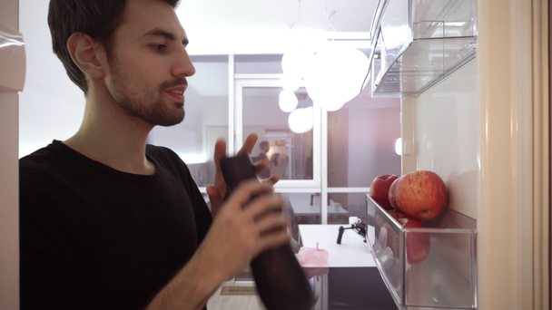 Zdravé stravování, jídlo a dieta koncept - tmavovlasý mladý muž otevření lednice doma kuchyně, pití a uvedení zpět černé sportovní láhev na chladničce police a odchodu - Záběry, video