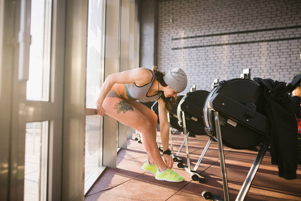 Beautiful sexy caucasian woman with a tattoo knots laces on green stems sitting to the will of a large window with sunlight in the gym - Photo, Image