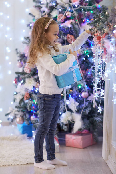 Szczęśliwa ładna dziewczyna z pudełkami na prezenty stoi w pobliżu choinki w pokoju domu. Wesołych Świąt i Nowego Roku. - Zdjęcie, obraz