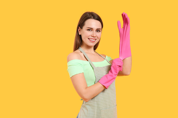 Young housewife putting on rubber gloves against color background - Photo, Image