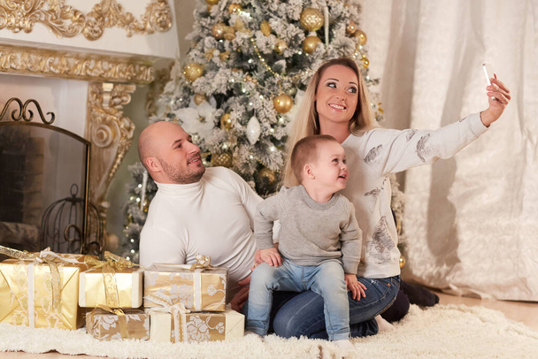 Happy family with a mobile phone and gift boxes sit near a Christmas tree in the house.  Merry Christmas and Happy New Year. - Foto, Imagen