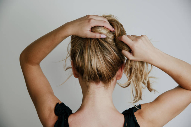 Rear view of a woman arranging her hair. Blond hair, tying hair in a bun. Woman on a light gray background. The concept of hairdressing, hair styling. - Фото, зображення