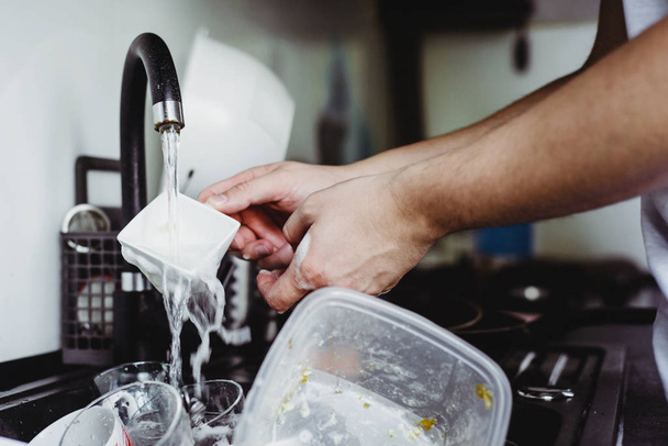 Чоловік чистить чашку після кави з шахраєм. Брудний посуд у раковині. Немає посудомийної машини. Концепція догляду за посудом на кухні. Кухонна раковина повна брудного посуду
. - Фото, зображення