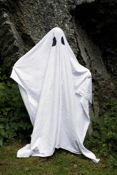 A spooky white ghost covered by a sheet with slits over the eyes standing in front of a rock. - Фото, изображение