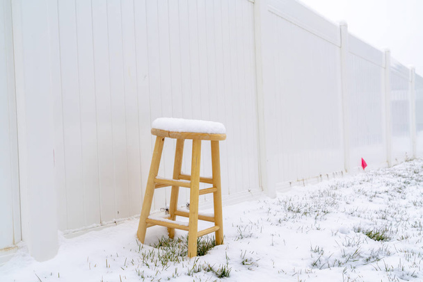 Wooden stool on snow covered ground in winter against white wood fence - 写真・画像