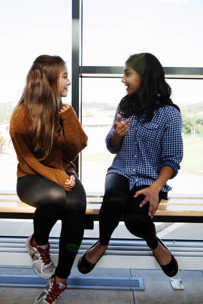 Two teenage schoolgirls sitting chatting in corridor - Photo, image