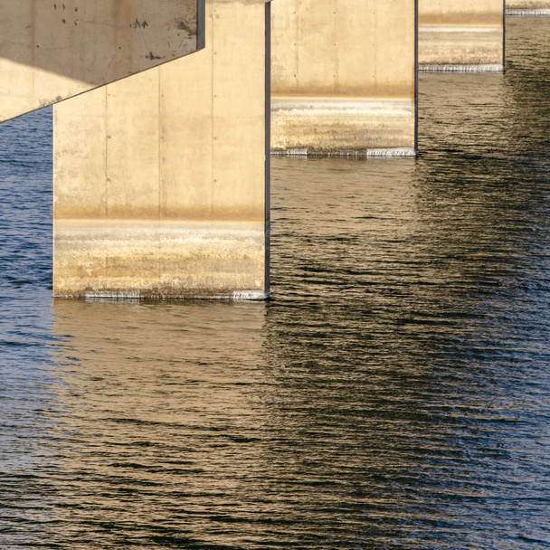 Vistas cuadradas debajo de un puente de vigas con pilares contra el agua del lago iluminada por el sol
 - Foto, Imagen